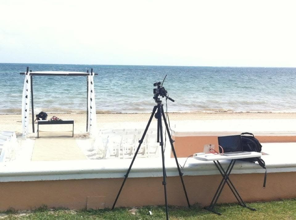 A camera on a tripod and a laptop on a folding table, set up facing a beach ceremony arch and rows of white chairs by the sea.