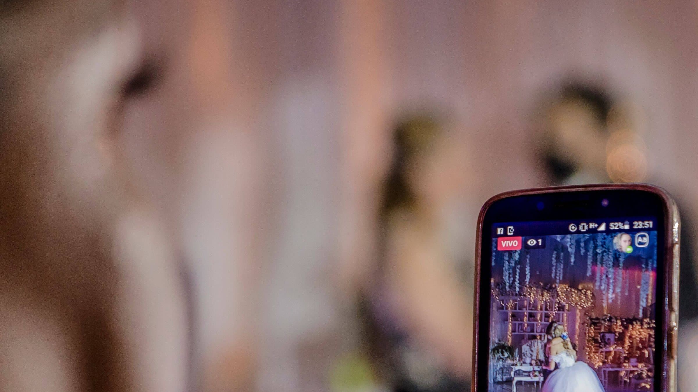 A guest holding up a phone at an evening reception, with the ceremony streaming live on the screen.
