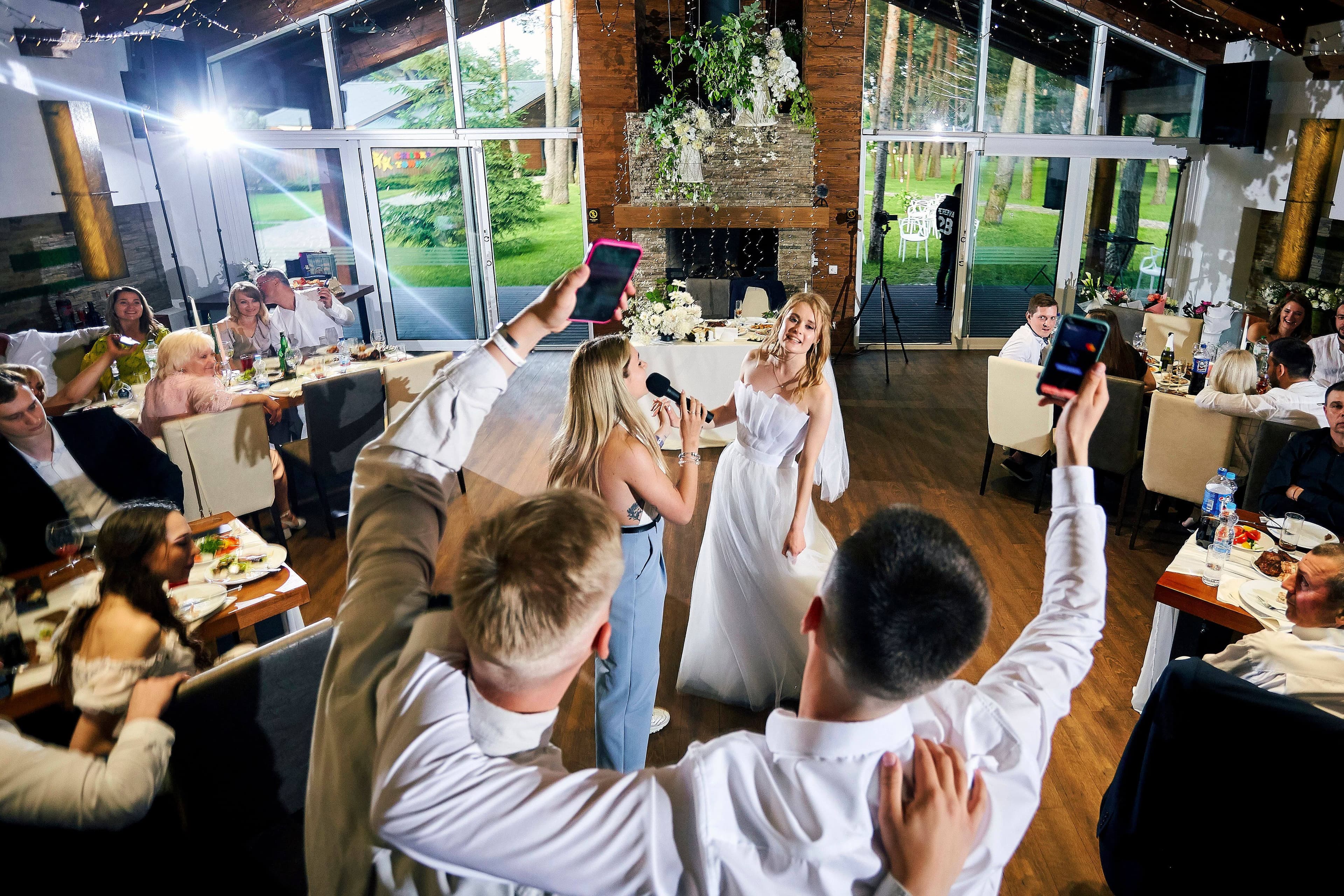 A reception in a glass-walled venue overlooking pine woods, with guests holding up their phones to share the moment.